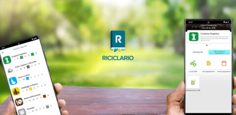 Due mani che reggono due cellulari con l'app Riciclario e sullo sfondo un paesaggio verdeggiante sfocato