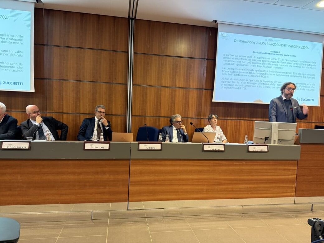 Uno speaker che parla da un palco mentre altre quattro persone sono sedute a un tavolo con microfoni