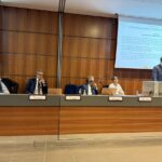 Uno speaker che parla da un palco mentre altre quattro persone sono sedute a un tavolo con microfoni