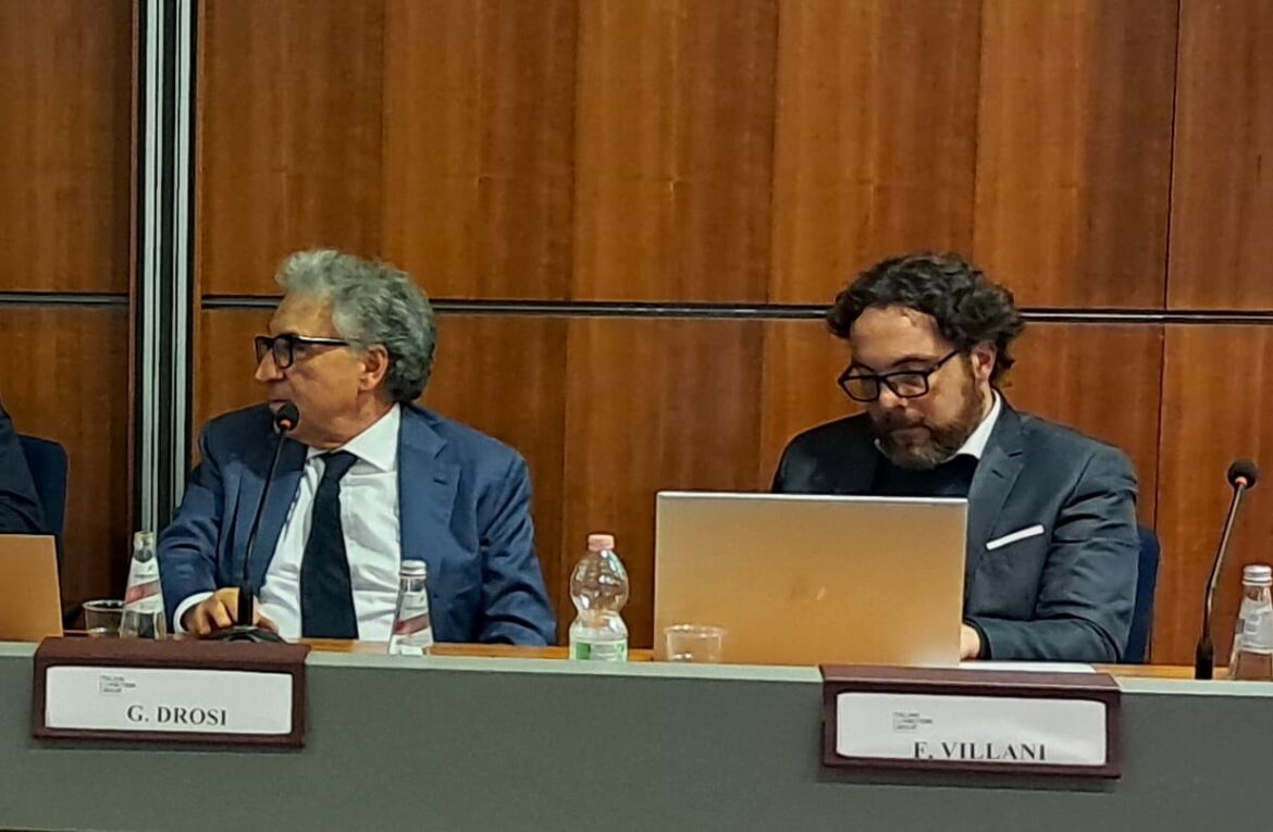 L'immagine di due persone sedute al tavolo di un convegno con i cartellini recanti i rispettivi nomi e un computer aperto davanti a uno dei due