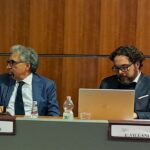 L'immagine di due persone sedute al tavolo di un convegno con i cartellini recanti i rispettivi nomi e un computer aperto davanti a uno dei due