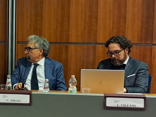 L'immagine di due persone sedute al tavolo di un convegno con i cartellini recanti i rispettivi nomi e un computer aperto davanti a uno dei due