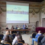 Persone sedute che osservano quattro relatori a un tavolo, uno di questi è in piedi con un microfono e sta parlando,