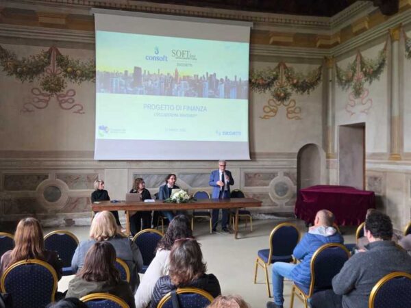 Persone sedute che osservano quattro relatori a un tavolo, uno di questi è in piedi con un microfono e sta parlando,