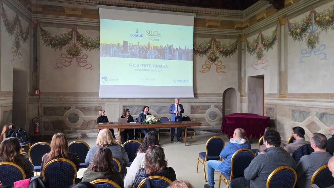 Persone sedute che osservano quattro relatori a un tavolo, uno di questi è in piedi con un microfono e sta parlando,