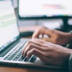 Le mani di una persona che digita su un portatile e sfocato sullo sfondo il monitor di un computer
